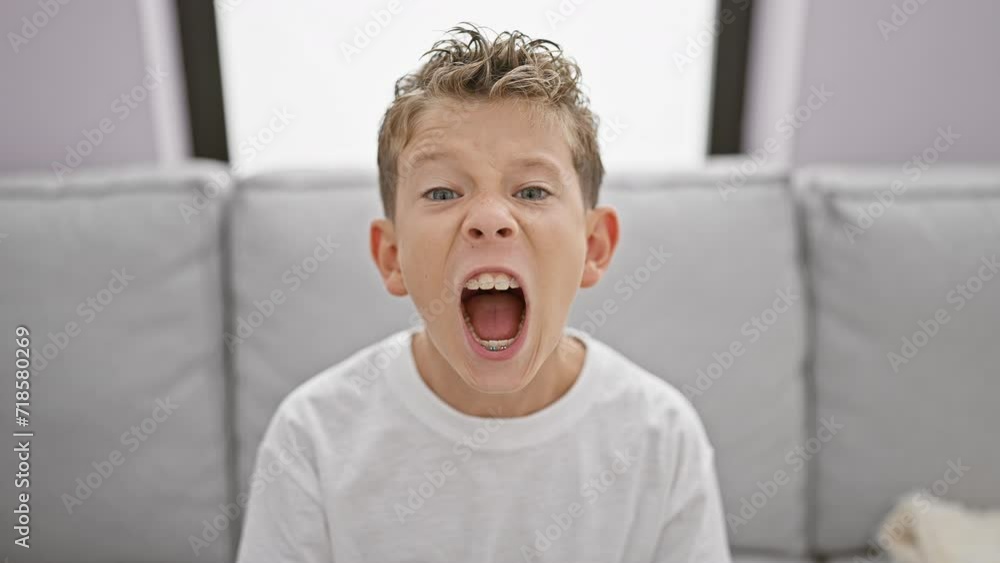 Adorable blond boy sitting on sofa at home, expressing loud frustration and anger Adorable blond boy sitting on sofa at home, expressing loud frustration and anger