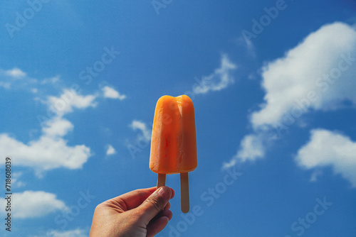 Hand holding an orange popsicle with the sea in the blue sky background. Delicious orange stick ice cream. Refreshing fruit ice creams against a beach scene