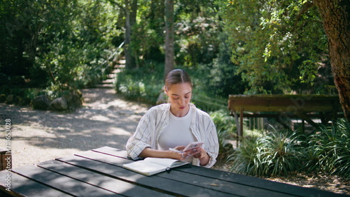 Happy woman reading message on cellphone sitting park table with book close up.