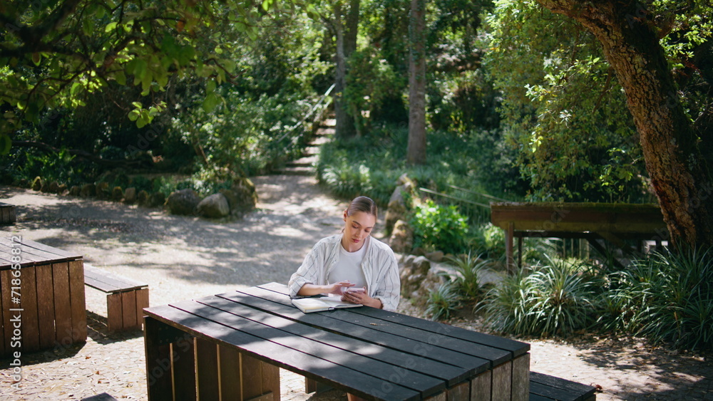 Fototapeta premium Woman sitting park table park with smartphone writing information into notebook