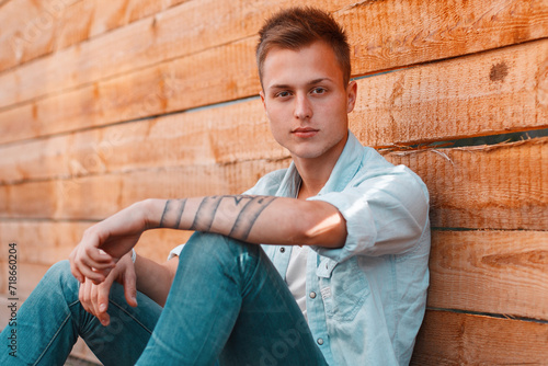 Stylish handsome young man with a hairstyle in a denim shirt with jeans sits near a wooden wall on the street