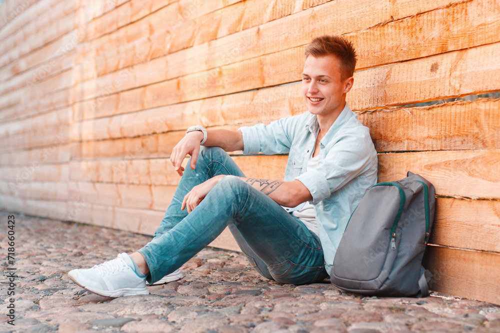 Happy handsome student man with a smile with a hairstyle in fashionable denim clothes with a shirt and jeans with a backpack sits on the ground near a wooden background. Traveler guy