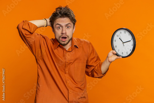Caucasian man with anxiety checking time on clock, running late to work, being in delay, deadline. Young guy looking at hour, minutes, worrying to be punctual isolated on studio orange background photo