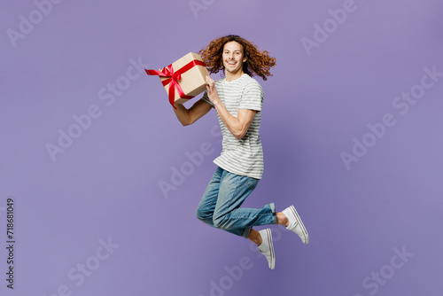 Full body young man wears grey striped t-shirt casual clothes hold in hand present box with gift ribbon bow jump high look aside isolated on plain pastel purple background studio. Lifestyle concept. photo