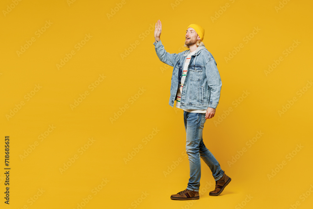 Full body side profile view smiling fun happy young man he wears denim shirt hoody beanie hat casual clothes waving hand walk go isolated on plain yellow background studio portrait. Lifestyle concept. Full body side profile view smiling fun happy young man he wears denim shirt hoody beanie hat casual clothes waving hand walk go isolated on plain yellow background studio portrait. Lifestyle concept.