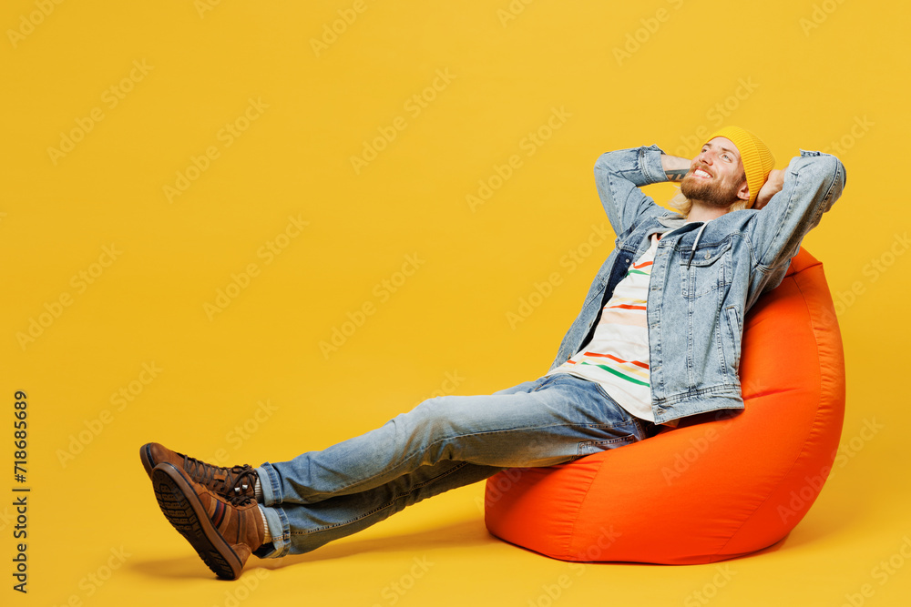 Full body side view minded young man wears denim shirt hoody beanie hat casual clothes sit in bag chair hold hands behind neck isolated on plain yellow background studio portrait. Lifestyle concept Full body side view minded young man wears denim shirt hoody beanie hat casual clothes sit in bag chair hold hands behind neck isolated on plain yellow background studio portrait. Lifestyle concept