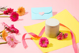 A box of unlabeled scented candles is displayed with gift wrap, ribbon, an envelope, gerberas and carnations on a pastel pink background. Close up.