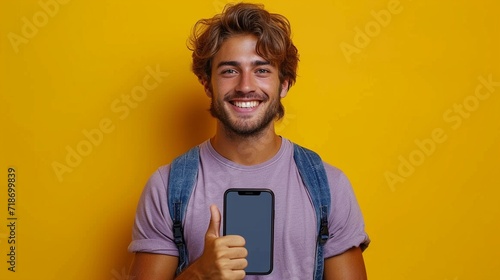 Explore a lifestyle moment as a smiling young man, donned in a purple t-shirt, proudly exhibits a smartphone with a big blank screen and gives a thumbs-up on a plain yellow background. photo