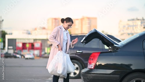 A tired young woman has done her shopping after work and loads packages into her car in the parking lot of a shopping center