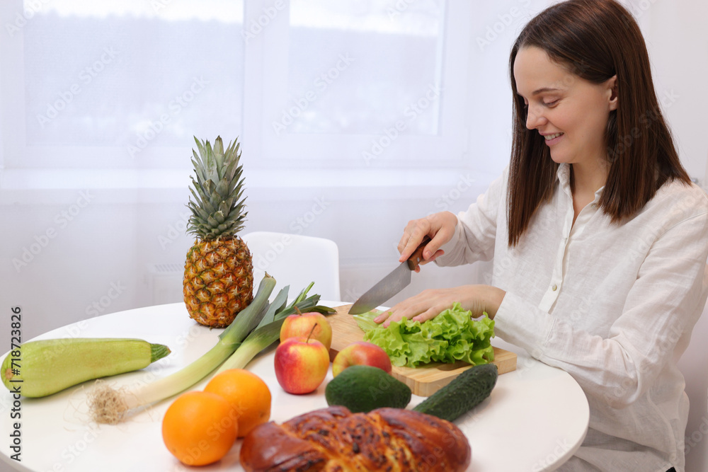 Pretty smiling Caucasian brown haired woman in white shirt sitting at kitchen table with fruit and vegetables cutting green lettuce leaves for healthy salad