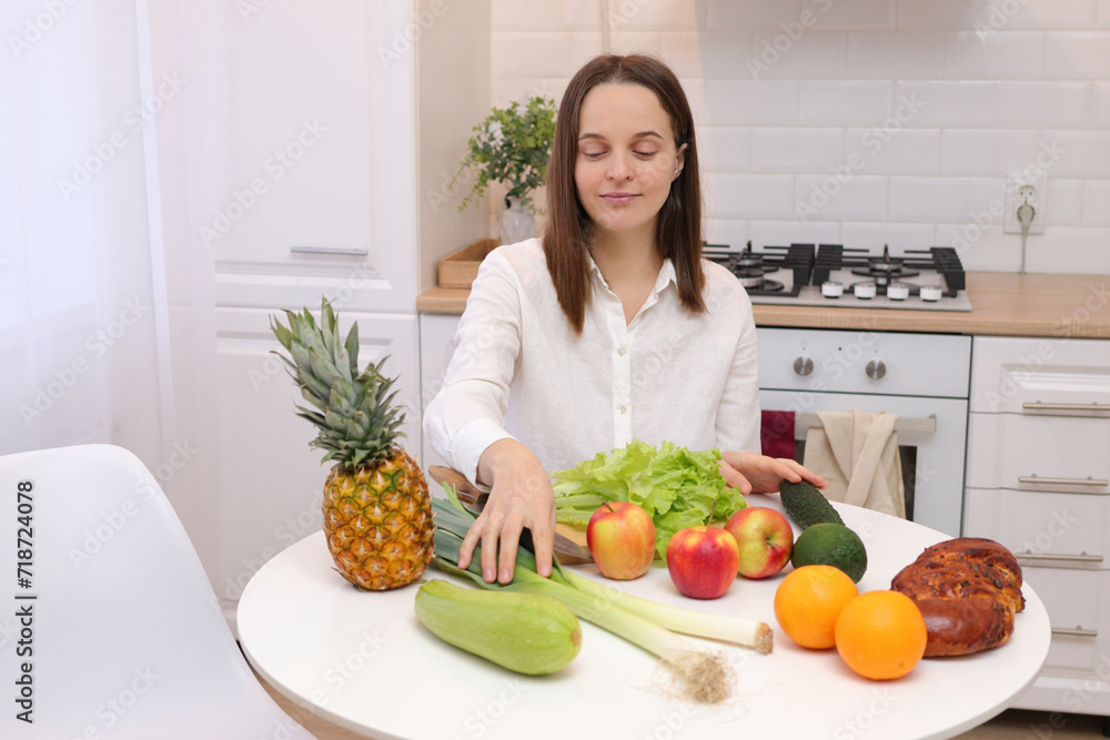 Healthy lifestyle. Raw vegan. Domestic kitchen. Nutritious dinner. Caucasian brown haired woman sitting at kitchen table with fruit and vegetables cooking organic meals at home