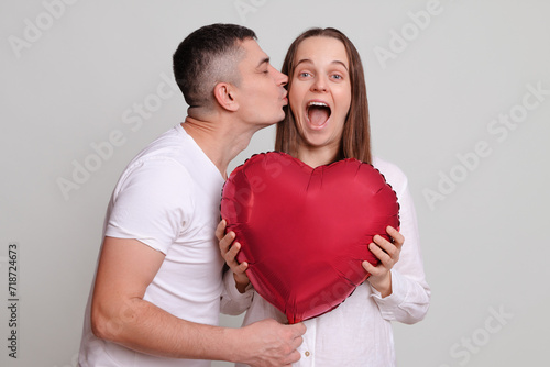 Loving husband kissing amazed excited wife on Valentines Day celebrating together Caucasian man and woman holding heart shaped air balloon isolated over gray background