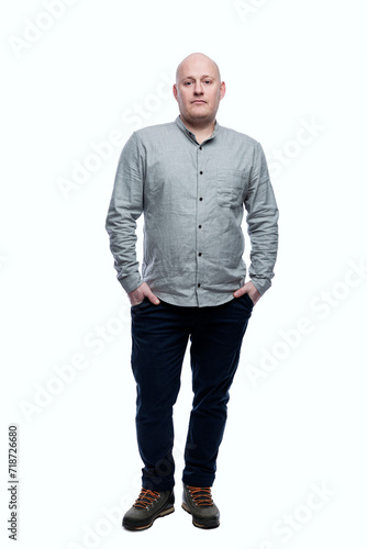 Serious bald adult man stands with his hands in his pockets. Wearing a gray shirt and blue jeans. Full height. Isolated on a white background. Vertical.