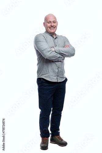 Laughing bald adult man stands with his arms crossed. A 40-year-old guy is wearing a gray shirt and dark blue jeans. Isolated on a white background. Full height. Vertical.