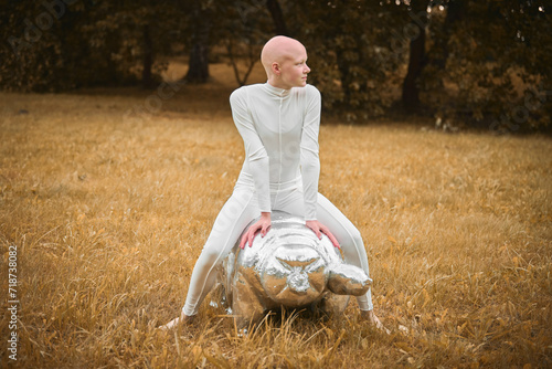 Young hairless girl with alopecia in white cloth sits on tardigrade figure in fall park, surreal scene with bald teenage girl confidently embraces her unique beauty photo