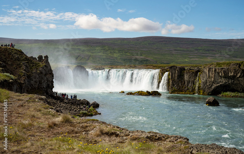 tiny tourists view the majestic Godafoss waterfall on the Skjalfandafljot river in northern Iceland with a beautiful green hillside in the background and wildflowers in the foreground