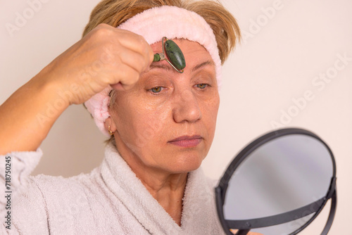 elder woman granny is using gua sha and jade roller for facial massage. anti wrinkle aging,rejuvenation procedures. portrait of old lady, with headband on head and mirror in hand.beauty skin care