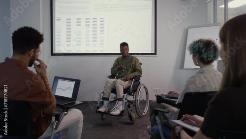 Full shot of young African American wheelchaired man pointing at graph on projector screen while making business presentation to diverse colleagues in office photo