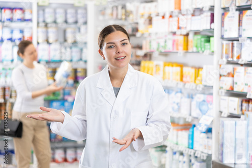 At pharmacy, smiling woman specialist stands in hall with open display and gestures to invite consultation about therapeutic cosmetics