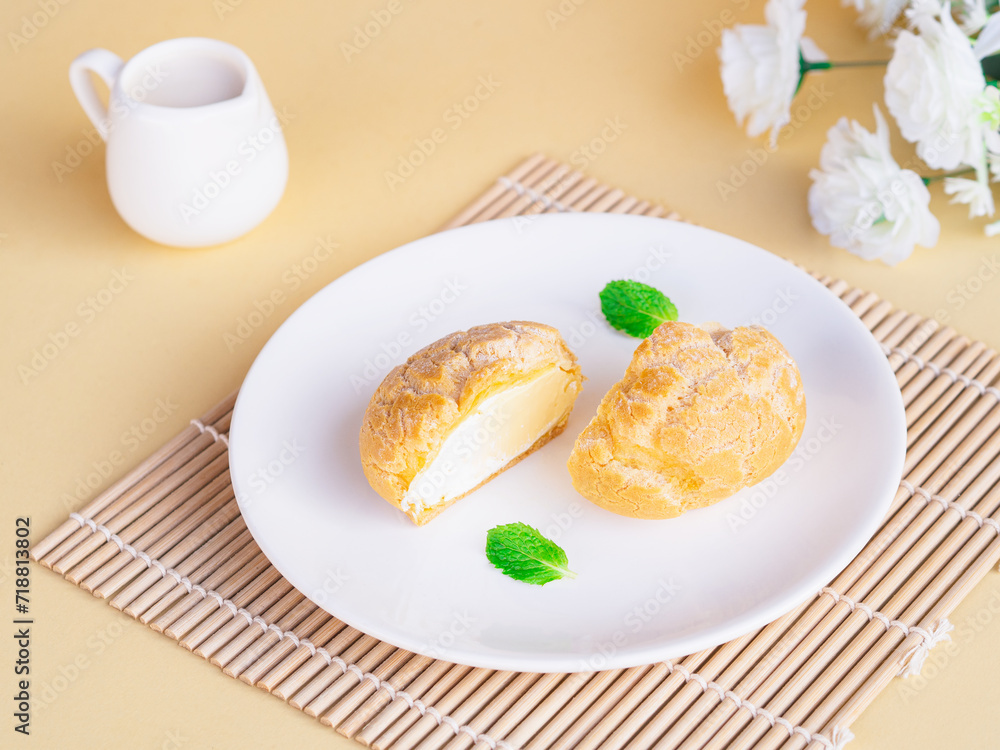 Half cut soft choux cream puff custard flavor in white plate on beige background. Baked Goods style and Minimalist product concept Half cut soft choux cream puff custard flavor in white plate on beige background. Baked Goods style and Minimalist product concept