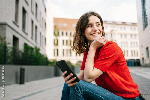 Young Student Sitting on Stone Steps in City, Laughing While Using Smartphone