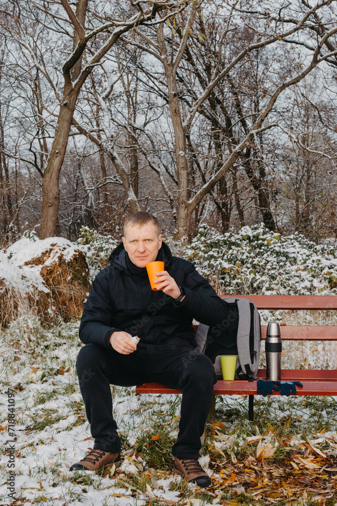 Fototapeta premium Winter Serenity: 40-Year-Old Man Enjoying Tea on Snow-Covered Bench in Rural Park. Immerse yourself in the tranquil beauty of winter as a 40-year-old man finds solace on a snowy bench in a rural park.