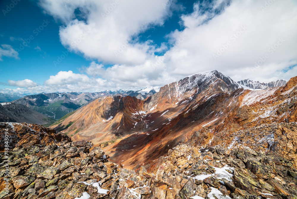 Scenic top view from stony precipice edge to multicolor big rocky ridge in freshly fallen snow and giant snow-capped mountain peak in low clouds. Snow-covered multi-color sharp rocks in huge mountains