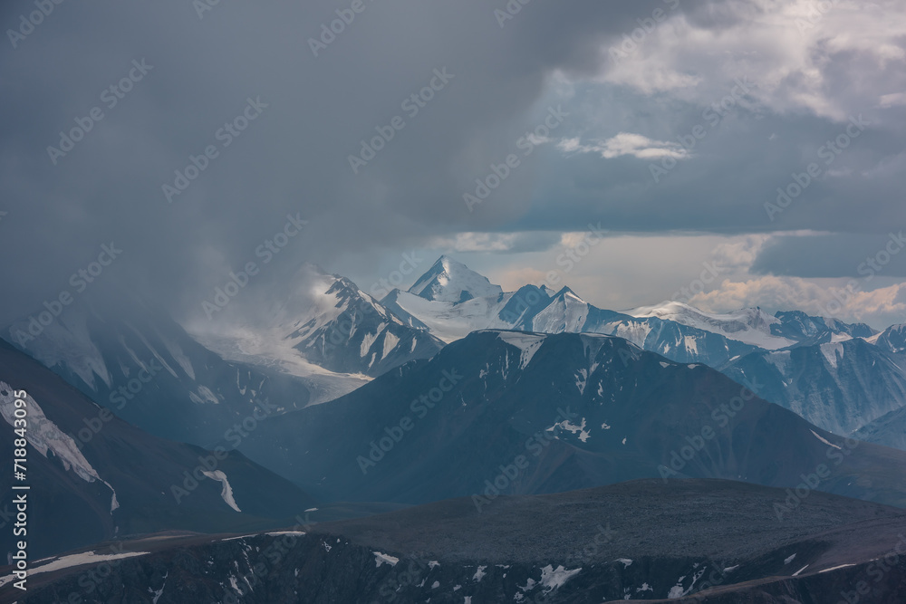 Dark atmospheric landscape with mountain silhouettes and large snow-capped peaked top in rainy low clouds. Dramatic alpine view to high mountains in gray cloudy sky. Big snowy mountain range in rain.