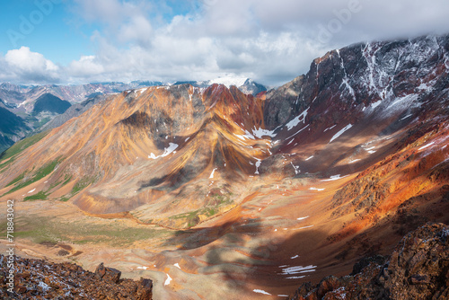 Top view from rocks to rounded multicolor valley with curved iron river and big sharp rocky ridge of red color. Colorful large mountain range in freshly fallen snow. Vivid high mountains in low clouds