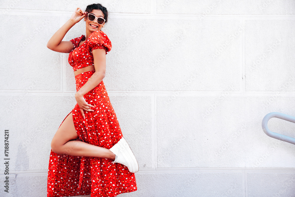 Young beautiful smiling hipster woman in trendy summer red dress. Carefree woman posing in the street near wall. Positive model outdoors in at sunny day. Cheerful and happy. In sunglasses Young beautiful smiling hipster woman in trendy summer red dress. Carefree woman posing in the street near wall. Positive model outdoors in at sunny day. Cheerful and happy. In sunglasses