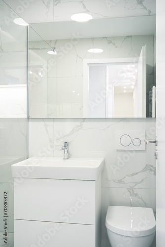 A Modern  bright bathroom with white marble walls  a glass shower  a sleek sink and cabinet  and a wall-mounted toilet.