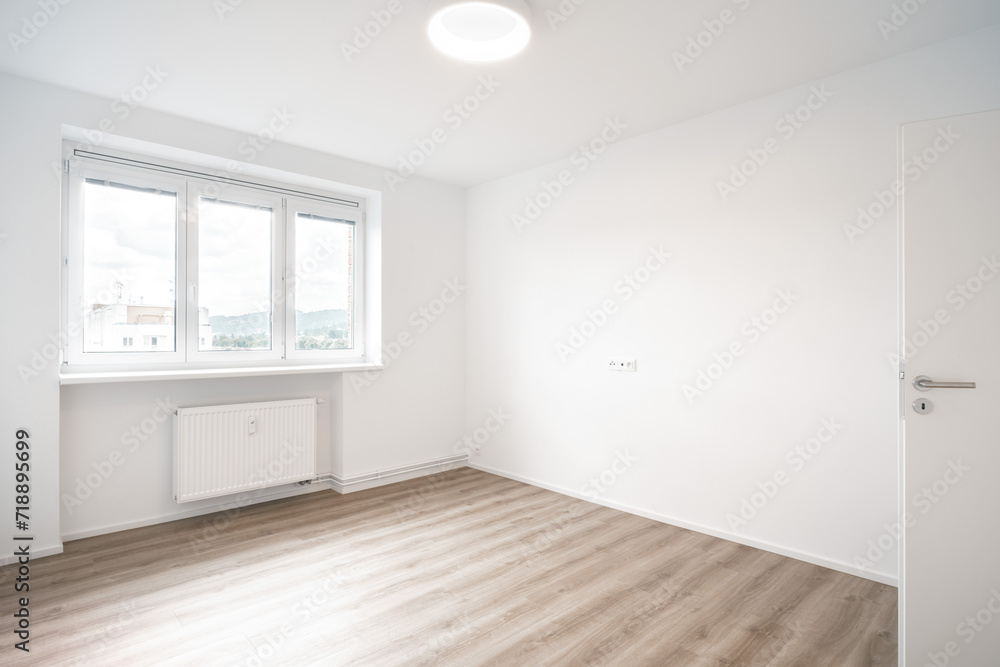 A Minimalist, bright room with white walls, a wooden floor, three windows, and an opened door. It’s clean and empty.