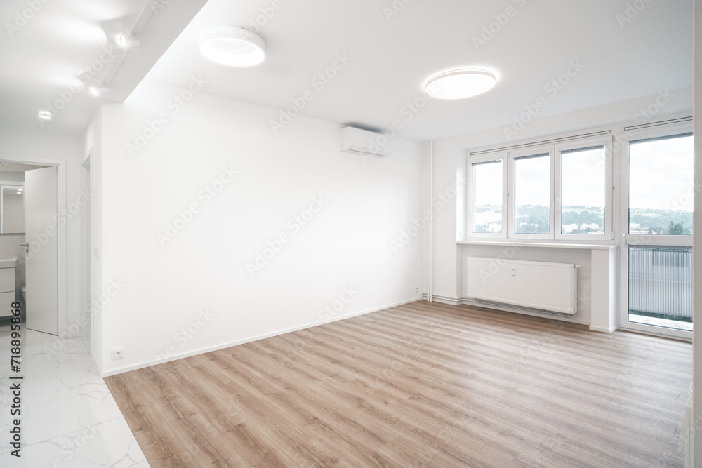 A Bright, spacious room with white walls, two ceiling lights, wooden flooring, and large windows. An open doorway leads to a tiled kitchen and bathroom area.