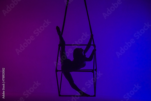 Silhouette of female aerial gymnast isolated on blue purple neon background. Female acrobat performing in the air on a cube, showing her flexibility.