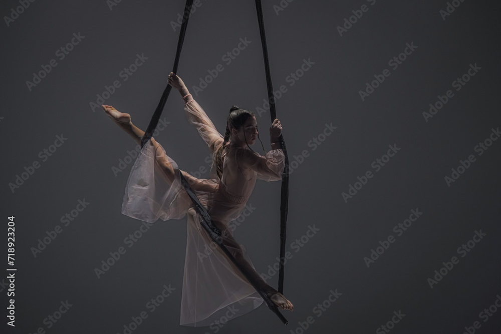 Girl aerial gymnast demonstrates stretching in twine on acrobatic trapeze. Acrobatic athlete performs hanging from a height in a dark studio.