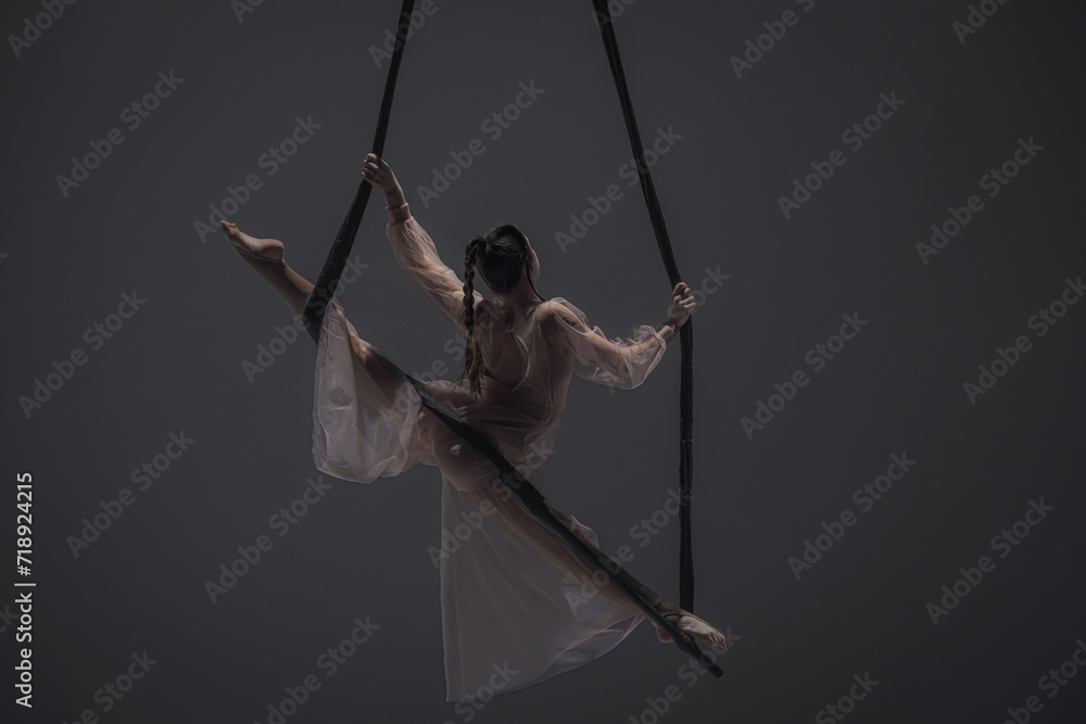 Girl aerial gymnast demonstrates stretching in twine on acrobatic trapeze. Acrobatic athlete performs hanging from a height in a dark studio. Girl aerial gymnast demonstrates stretching in twine on acrobatic trapeze. Acrobatic athlete performs hanging from a height in a dark studio.