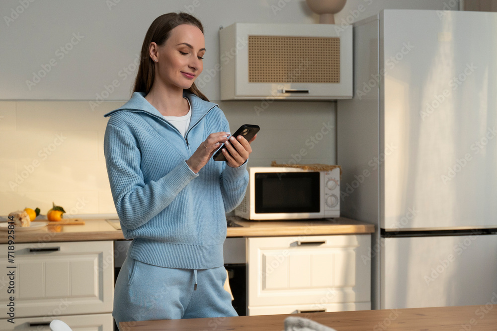 Young happy housewife dressed in a blue sweater, holding a mobile phone in her hands, looking for a recipe, standing alone in the bright kitchen of the house. Young happy housewife dressed in a blue sweater, holding a mobile phone in her hands, looking for a recipe, standing alone in the bright kitchen of the house.
