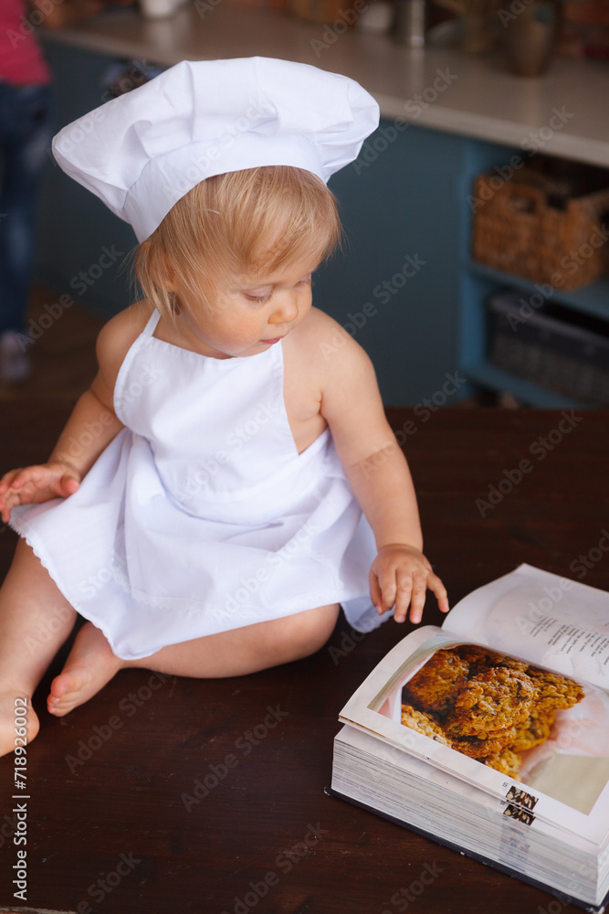 A baby girl, wearing a cooking hat and apron, is sitting on the table and reading a cooking book