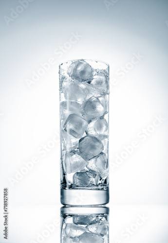 A tall transparent glass cup filled with natural ice cubes on a light background, backlit, studio shot.