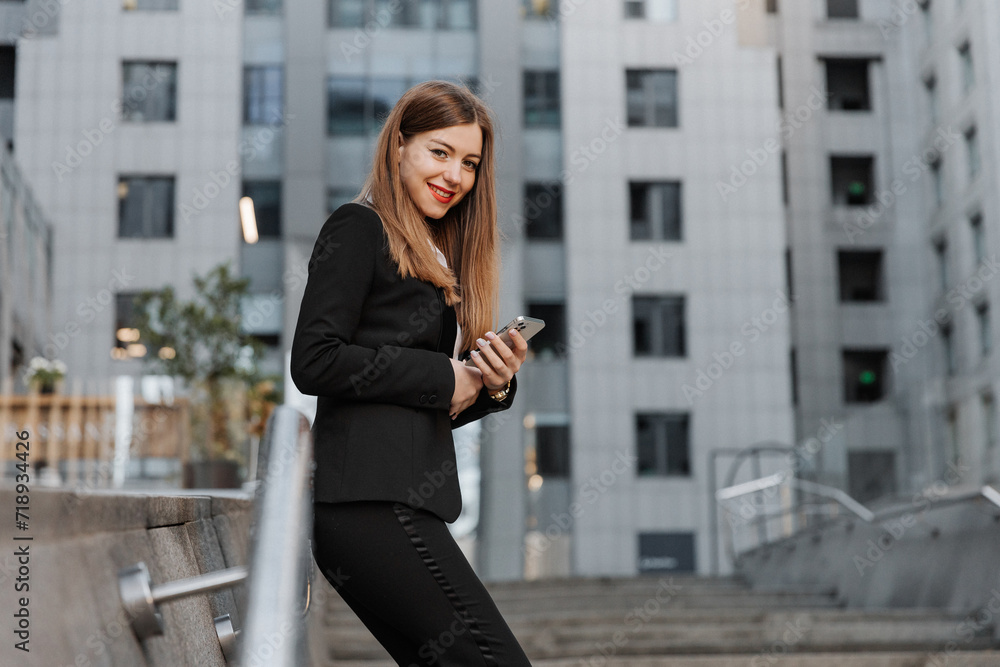 Young business woman wearing black dress code smiling answering message or chatting, using phone and looking at the camera