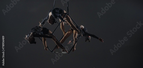 Trio of young female acrobats posing on a cube suspended at a height. Aerial gymnasts perform in studio against dark background and demonstrate flexibility and stretching The concept of a circus show