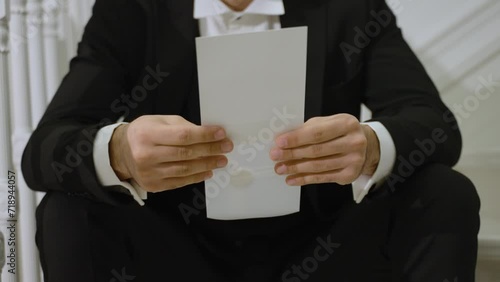 man in a black suit holding a white envelope. He is seated and focused on the content, possibly preparing to read a letter or an invitation. conveys a sense of anticipation and formality