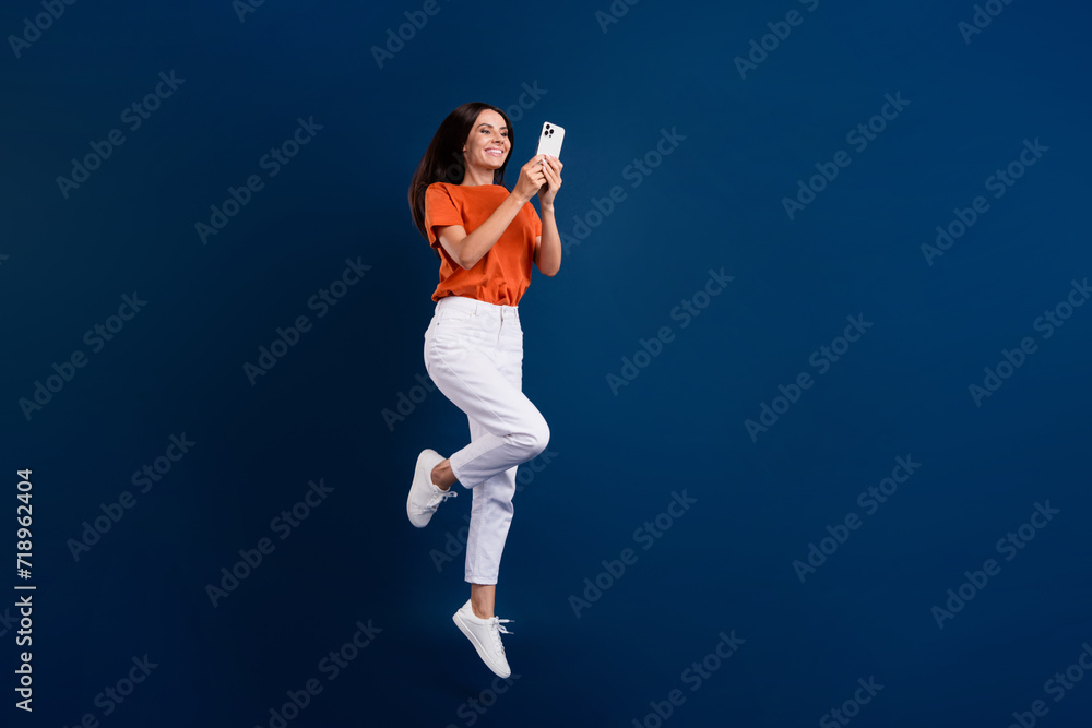 Full length photo of shiny cheerful lady wear orange t-shirt jumping chatting modern device empty space isolated dark blue color background