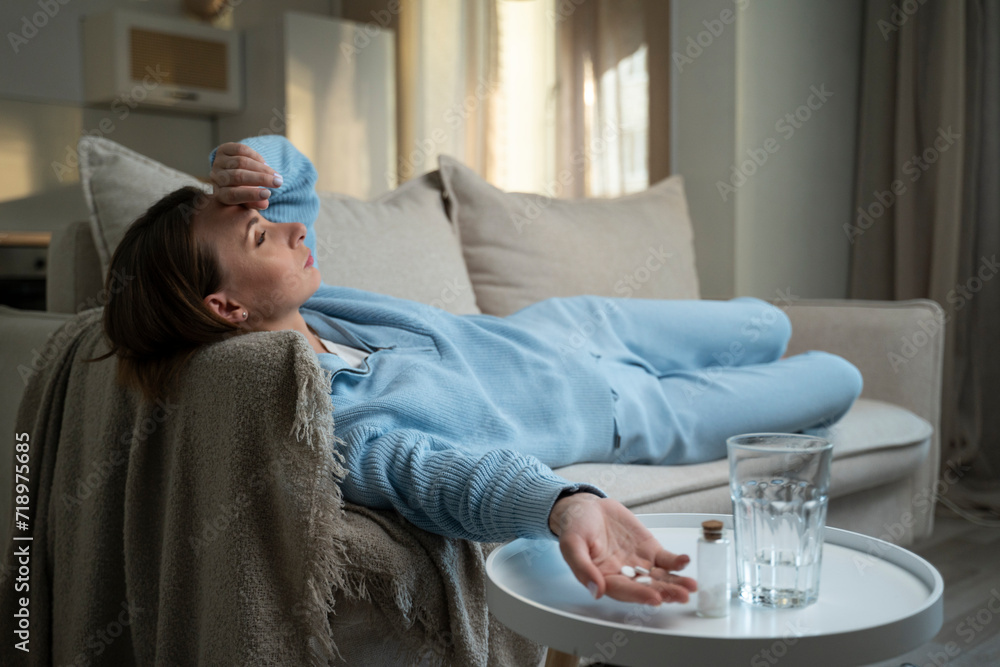 Young woman touches her forehead to check her body temperature, a girl has a headache when she lies at home on the couch with pills, drinks a glass of water and pills. Young woman touches her forehead to check her body temperature, a girl has a headache when she lies at home on the couch with pills, drinks a glass of water and pills.