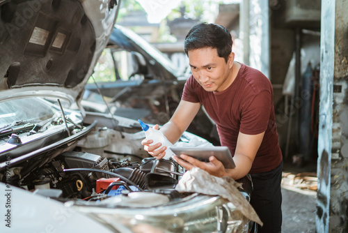 car mechanic using a laptop while holding a battery water bottle to maintain a car battery at a repair shop