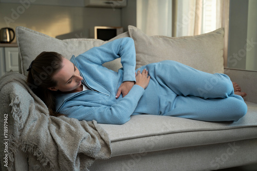 Sick, unhappy young brunette suffers from abdominal pain in everyday life, lies at home on the couch holding her stomach, experiences abdominal pain and discomfort, suffers from menstrual cramps.  © Grustock