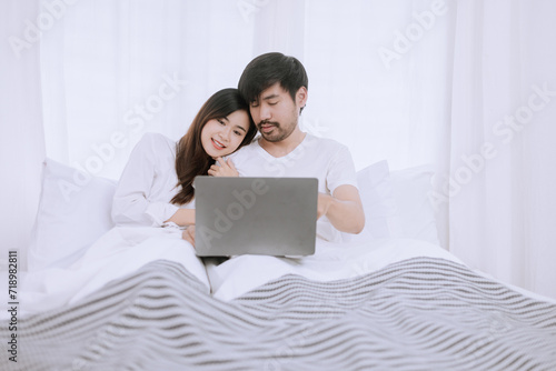 Happy asian man and woman using laptop sitting relaxed on a bed at home enjoy, Happy lover hug each other and smiling on the bed, Male and female couple concept.