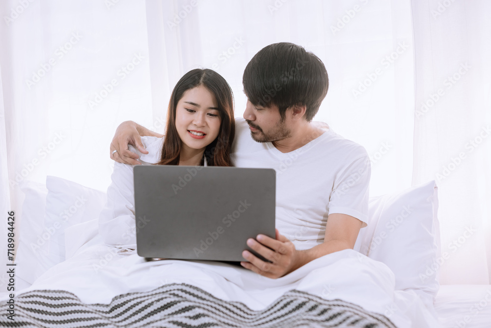 Happy asian man and woman using laptop sitting relaxed on a bed at home enjoy, Happy lover hug each other and smiling on the bed, Male and female couple concept.