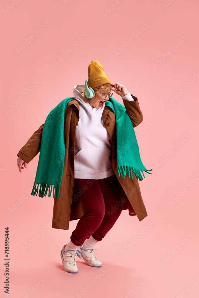 Charming, excited elderly woman in fashion outfit dancing while listening energetic music against pink background. Concept of active seniors in modern life, seniors using technology.