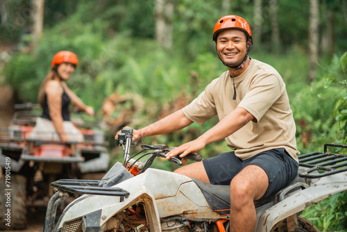 handsome asian man smiling to the camera while riding his atv with his beautiful partner at the background